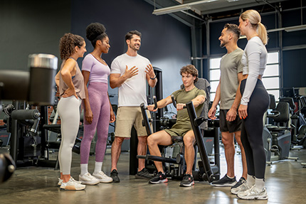 Ausbilder mit einer Gruppe von Fitnessstudierenden an einem Fitnessgerät. Junger Mann zeigt daran eine Übung.